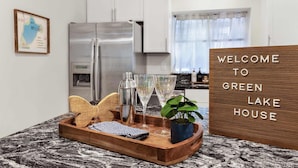 A kitchen with a stainless steel refrigerator, a wooden tray with a plant, glasses, and a cocktail shaker. A sign reads "WELCOME TO GREEN LAKE HOUSE".