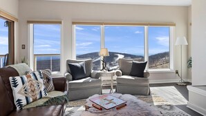 A living room with a brown leather sofa, two grey armchairs, a coffee table with a board game, and a view of a snowy landscape through large windows.
