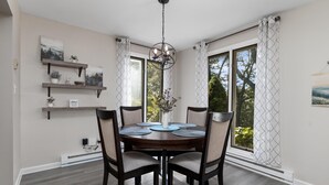 Ein Esszimmer mit einem runden Tisch, vier Stühlen und einer Deckenleuchte. Zwei Fenster mit Vorhängen und eine Wandregal mit Dekorationen.