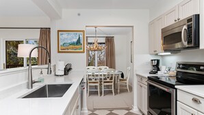 A modern kitchen with white cabinetry, a stainless steel sink, a built-in microwave, and an oven. There is a dining area with a table and chairs, and a chandelier hanging above.