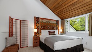 A bedroom with a large bed, a wooden ceiling, a window with a view of trees, a traditional Japanese screen, and a bedside table with lamps.