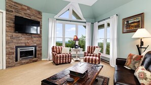 Un salón con chimenea de piedra, dos sillones a cuadros, un sofá de cuero oscuro, una mesa de centro de madera y ventanas grandes con cortinas.