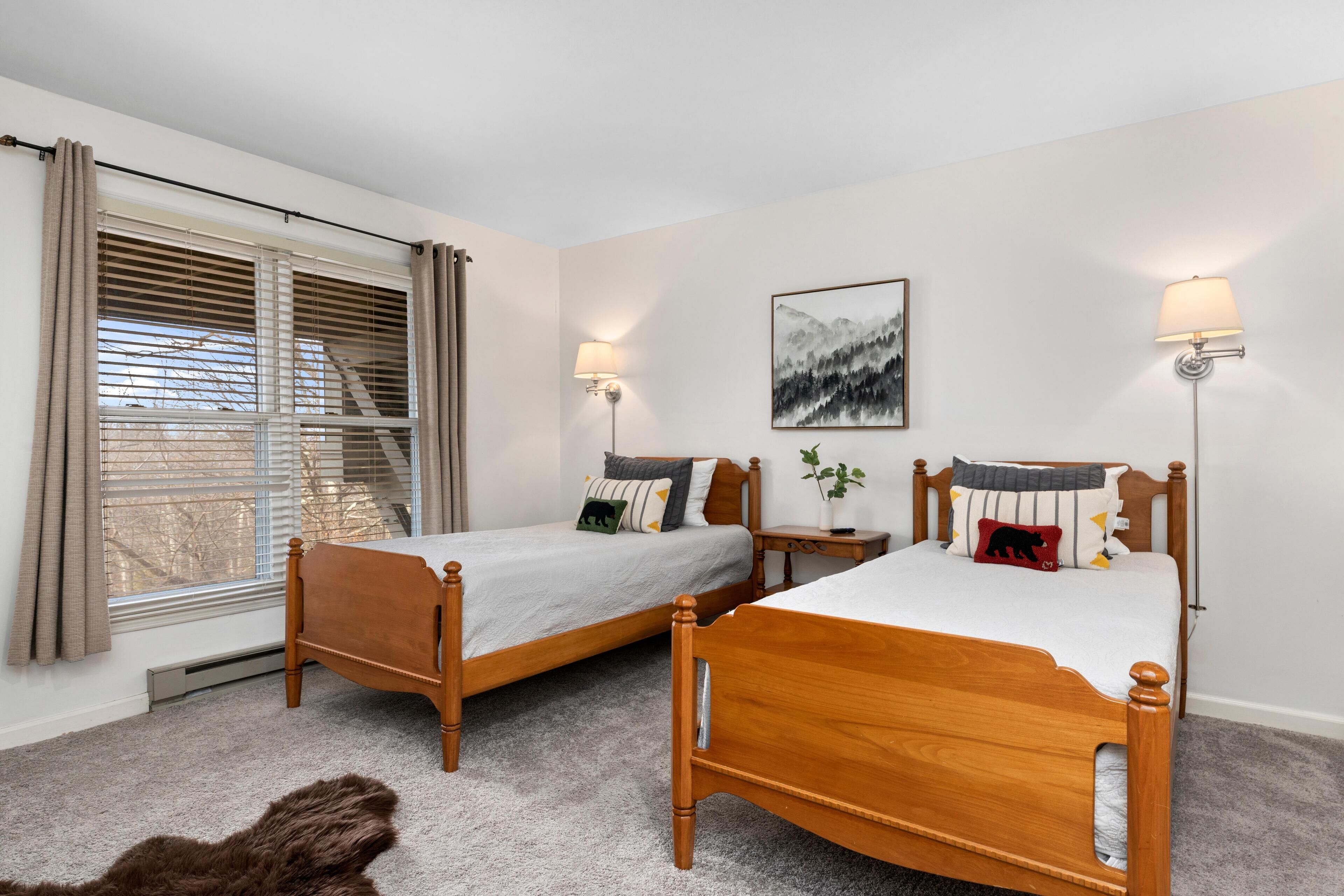 Een slaapkamer met twee houten bedden, een groot raam met jaloezieën, een wandlamp en een lijst met een afbeelding.