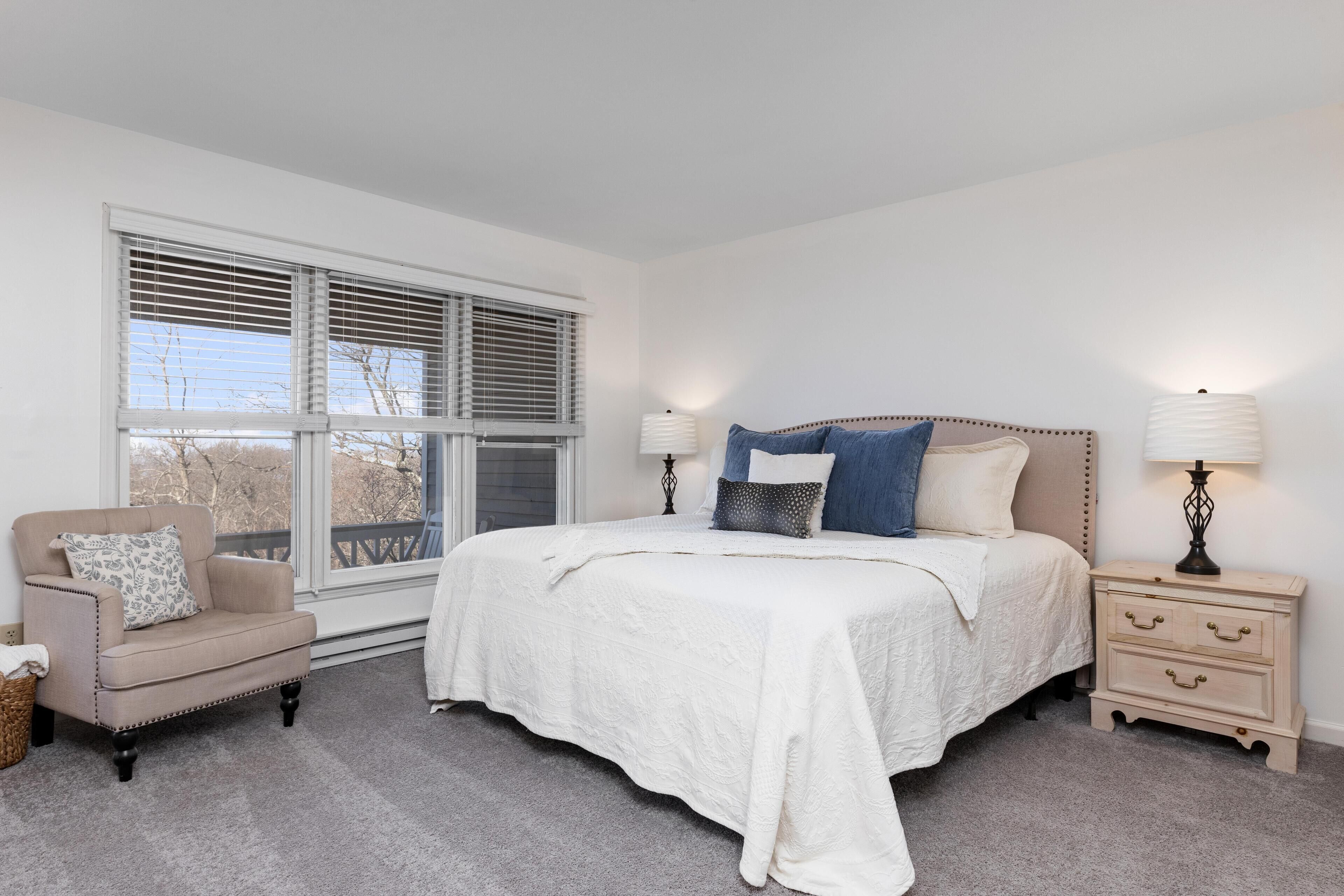 A bedroom with a large bed, two bedside tables, a chair, and a window with blinds.