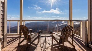 A wooden deck with two Adirondack chairs and a small round table, overlooking a mountainous landscape.