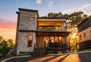 A two-story modern house with a stone facade, a balcony with seating, and a large window.