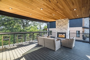 An outdoor deck with a stone fireplace, wicker furniture, and a view of greenery. - Serene Cabin Retreat・Rooftop Deck・Hot Tub・Games (Sevierville)
