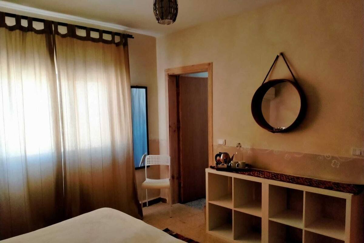 A hotel room with a bed, a small white chair, a mirror, and a shelf with a kettle and cups.