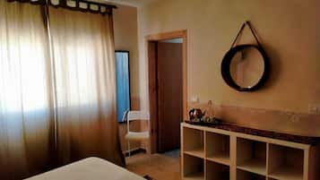 A hotel room with a bed, a small white chair, a mirror, and a shelf with a kettle and cups.