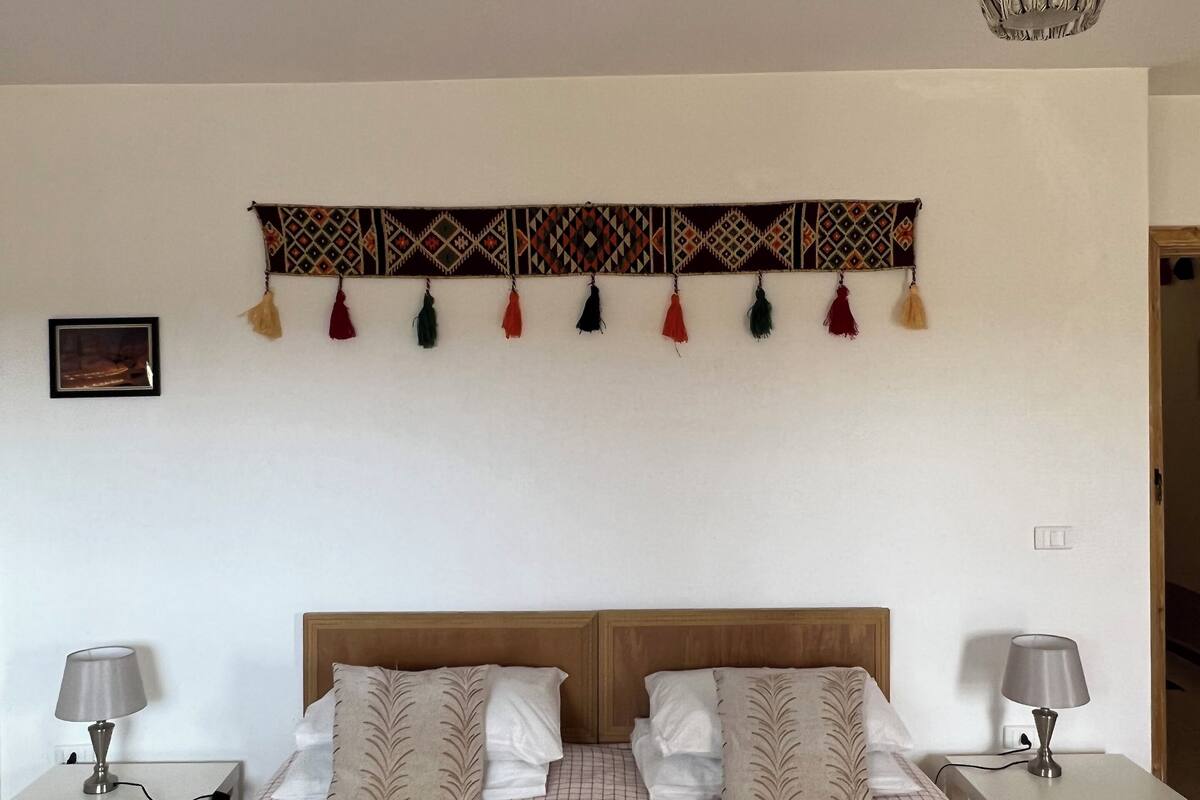 A bedroom with a wooden headboard, a patterned bedspread, two bedside tables with lamps, a wall hanging with tassels, and a hanging light fixture.