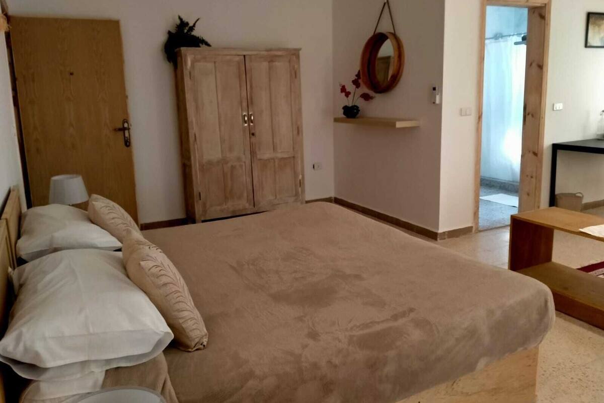 A bedroom with a large bed, a wooden wardrobe, a mirror on the wall, and a small table.