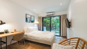 Habitación de hotel con cama, escritorio, silla, televisor y balcón con vista a follaje.
