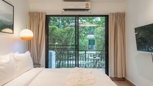 Habitación de hotel con cama, televisor, aire acondicionado, balcón con mesa y sillas, y cortinas.