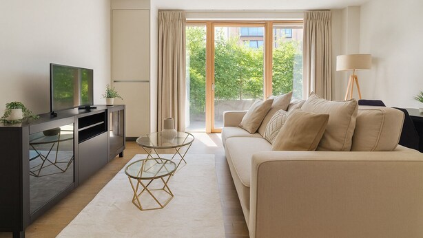 A modern living room with a sofa, a glass coffee table, a TV on a cabinet, and a view of greenery outside. - Modern 2-Bed flat in Mill hill Park (London)