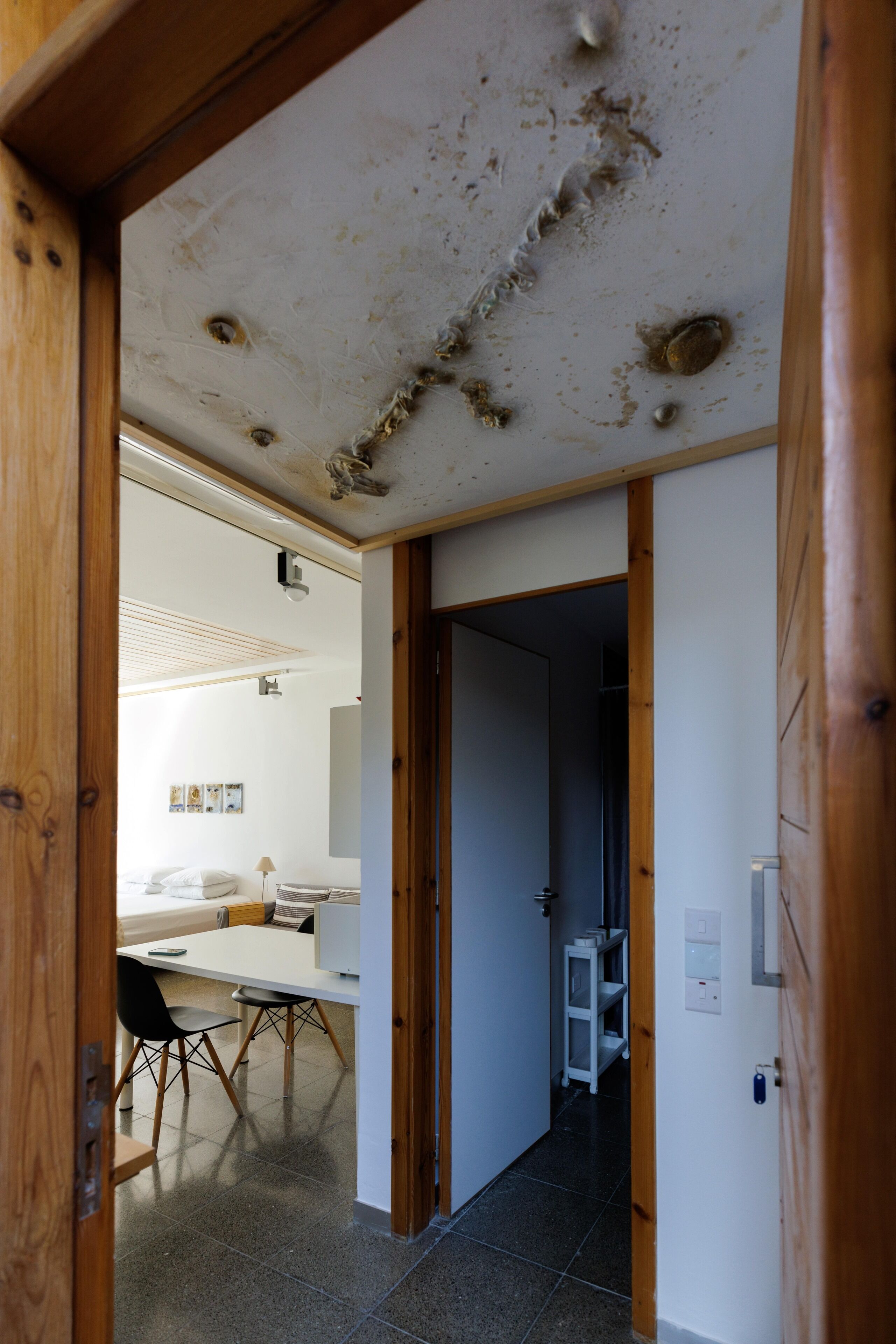 Un couloir mène à une salle à manger équipée d’une table et de chaises, un mur blanc orné d’une étagère et une porte.