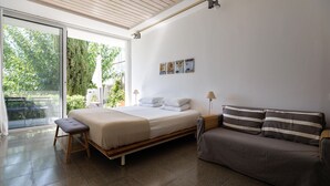 A modern bedroom with a bed, a bench, a sofa, and a view of greenery outside. - Eupnea | Studio Apartment, Art by PraXis Apartments (Limassol)