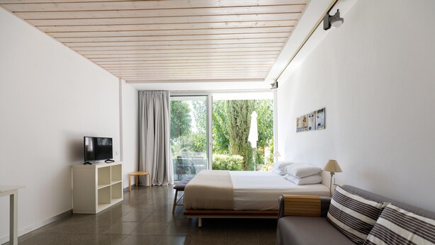 A modern bedroom with a bed, a sofa, a TV, and a balcony with a view of greenery. - Eupnea | Studio Apartment, Art by PraXis Apartments (Limassol)