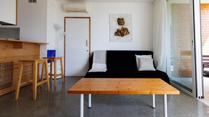 Una sala de estar moderna con una mesa de madera, dos taburetes de madera, un sofá negro con cojines blancos, una obra de arte colgada en la pared y un divisor de vidrio corredizo que da a un espacio exterior.