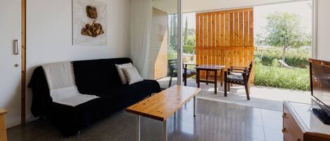 A modern living room with a black sofa, a wooden coffee table, and a TV on a wooden cabinet. There is a sliding glass door leading to an outdoor area with a wooden table and chairs.