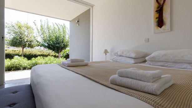 Una cama bien hecha con ropa de cama y toallas blancas, una lámpara de noche y vistas al exterior con vegetación.