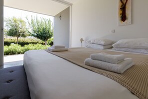 Un lit bien fait, avec du linge de lit et des serviettes blanches, une lampe de chevet et une vue sur la verdure à l’extérieur.