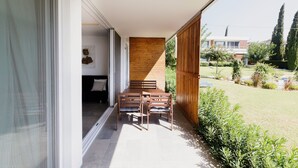 Un patio moderno con una mesa y sillas de madera, una puerta corrediza de vidrio que da a una habitación y vistas a una zona residencial con árboles y un edificio al fondo.