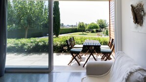 A modern living room with a sliding glass door leading to a patio with a dining set and a view of a well-maintained garden. - Euphonia | One-Bedroom Apartment, Art by PraXis Apartments (Limassol)