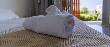 A neatly made bed with a rolled white towel, a textured beige bedspread, and a view of an outdoor area with greenery and a building in the background.