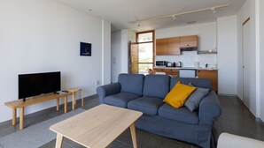 Un salon moderne avec un canapé, un téléviseur, une table en bois et un coin cuisine à l’arrière-plan.