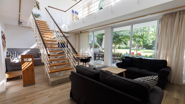 Een ruime woonkamer met een houten trap, zwarte banken, een houten salontafel en een grote schuifpui die uitkomt op een terras.