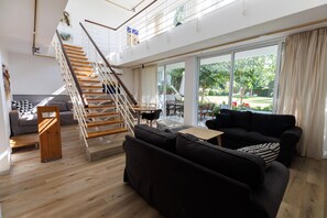 Un vaste espace de vie agrémenté d’un escalier en bois, de canapés noirs, d’une table basse en bois et d’une grande porte-fenêtre coulissante donnant sur une terrasse extérieure.