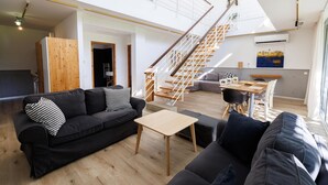 Een ruime woonkamer met een zwarte bank, een houten salontafel en een trap naar een hoger gelegen verdieping.