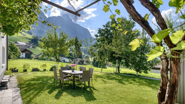 A patio with a table and chairs, surrounded by grass and trees, with a mountain view in the background. - Gartenferienwohnung Arlberg (Bludenz)