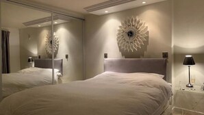 A hotel room with two beds, a large mirror, a decorative wall piece, and a bedside table with a lamp.