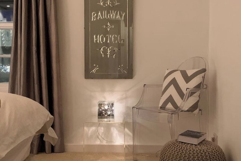 A hotel room with a bed, a chair with a patterned cushion, a small table with a book, and a wall-mounted sign that reads "RAILWAY HOTEL".