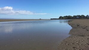 A beach with calm waters, a sandy shore, and a distant shoreline with vegetation and buildings.