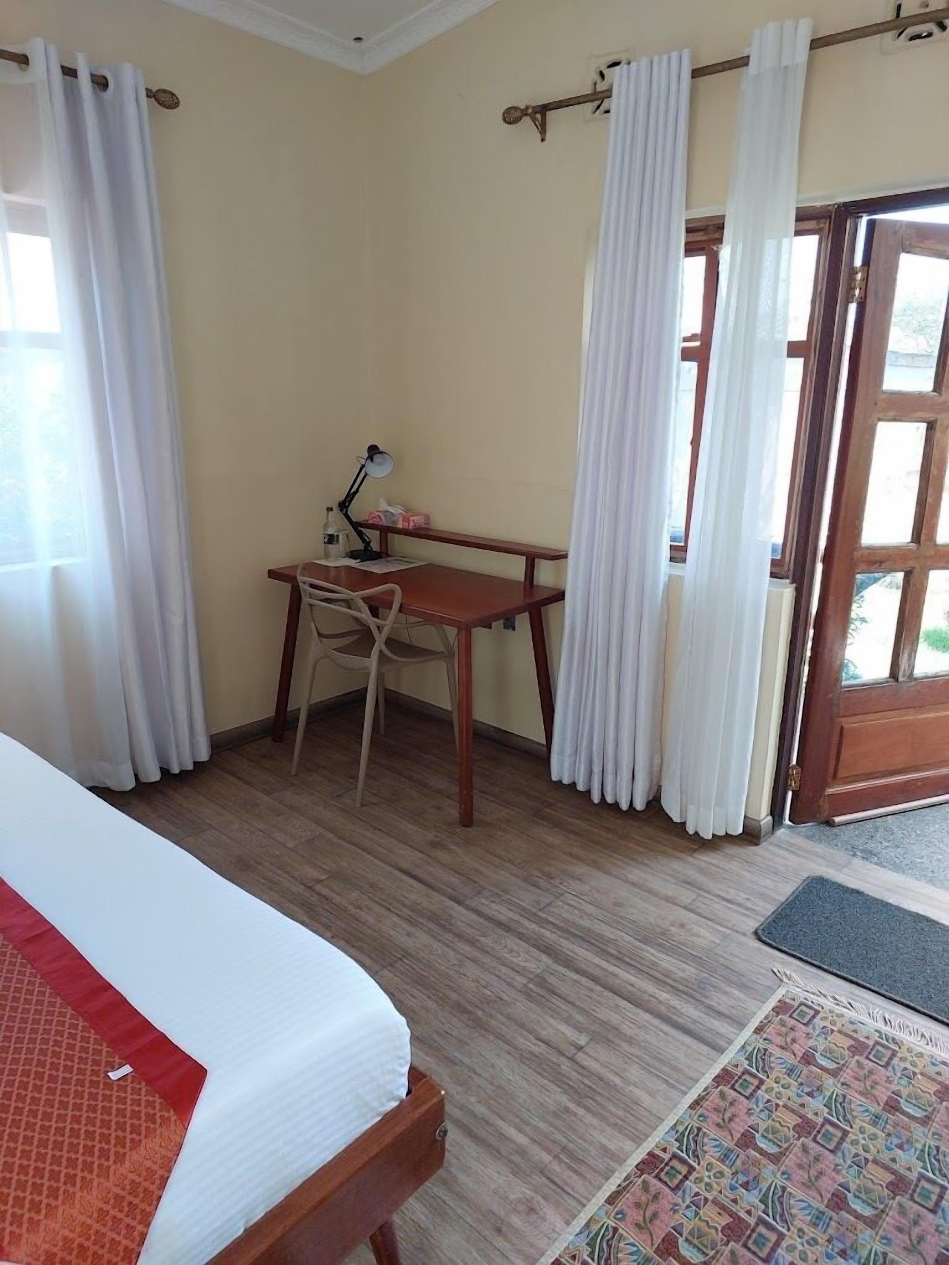 Una habitación con una cama, un escritorio y una silla. Hay una ventana con cortinas, una puerta y una alfombra en el suelo.