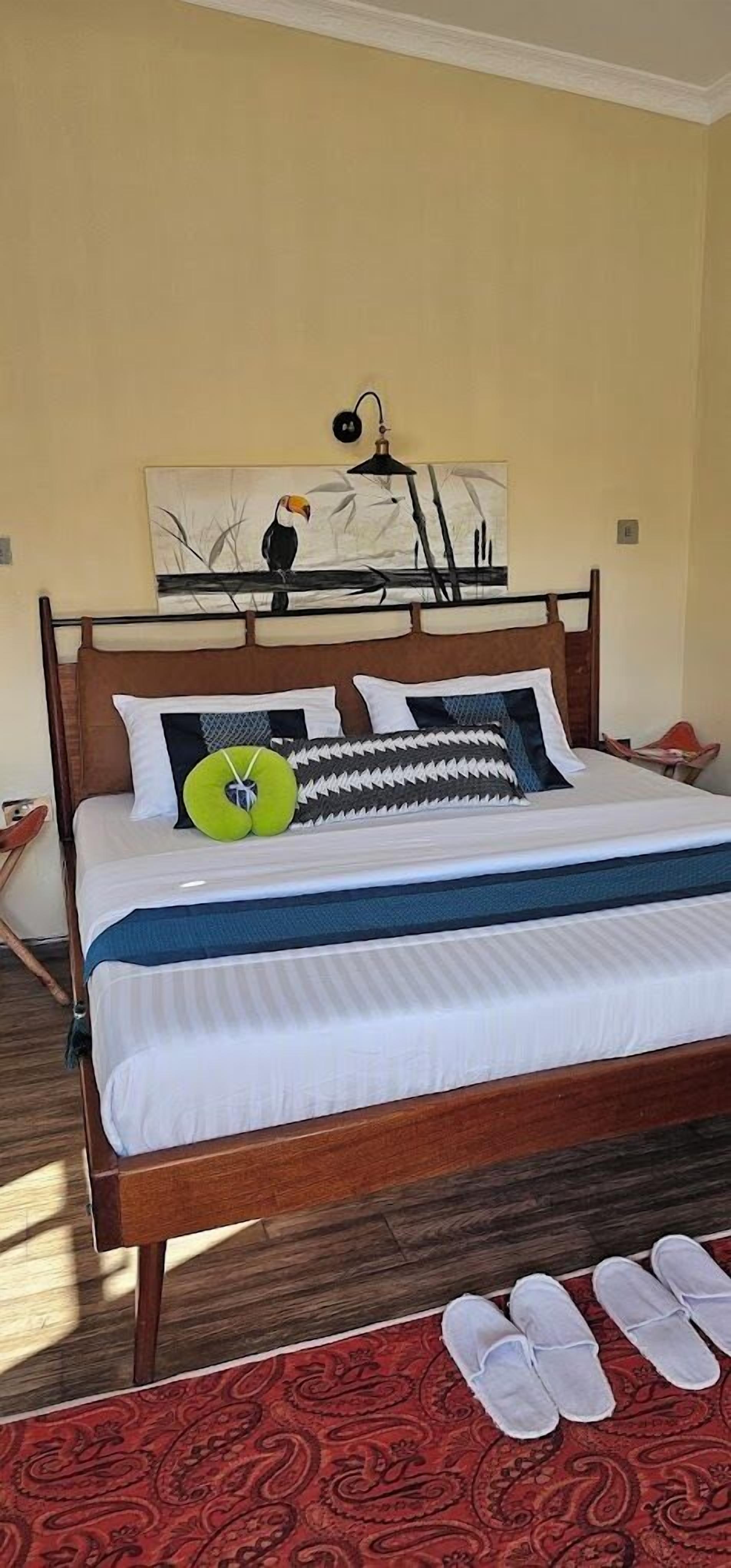 A neatly made bed with a headboard, a painting above it, and a pair of slippers on a patterned rug.