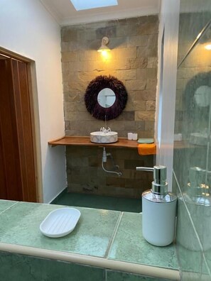Baño con pared de piedra, un espejo redondo, un lavamanos con dispensador de jabón y zona de ducha.