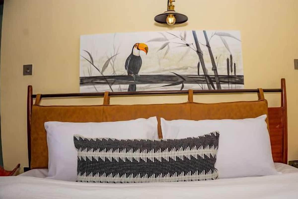 A bed with a wooden headboard, a white pillow, and a black and white patterned pillow. Above the bed, there is a painting of a toucan on a branch.