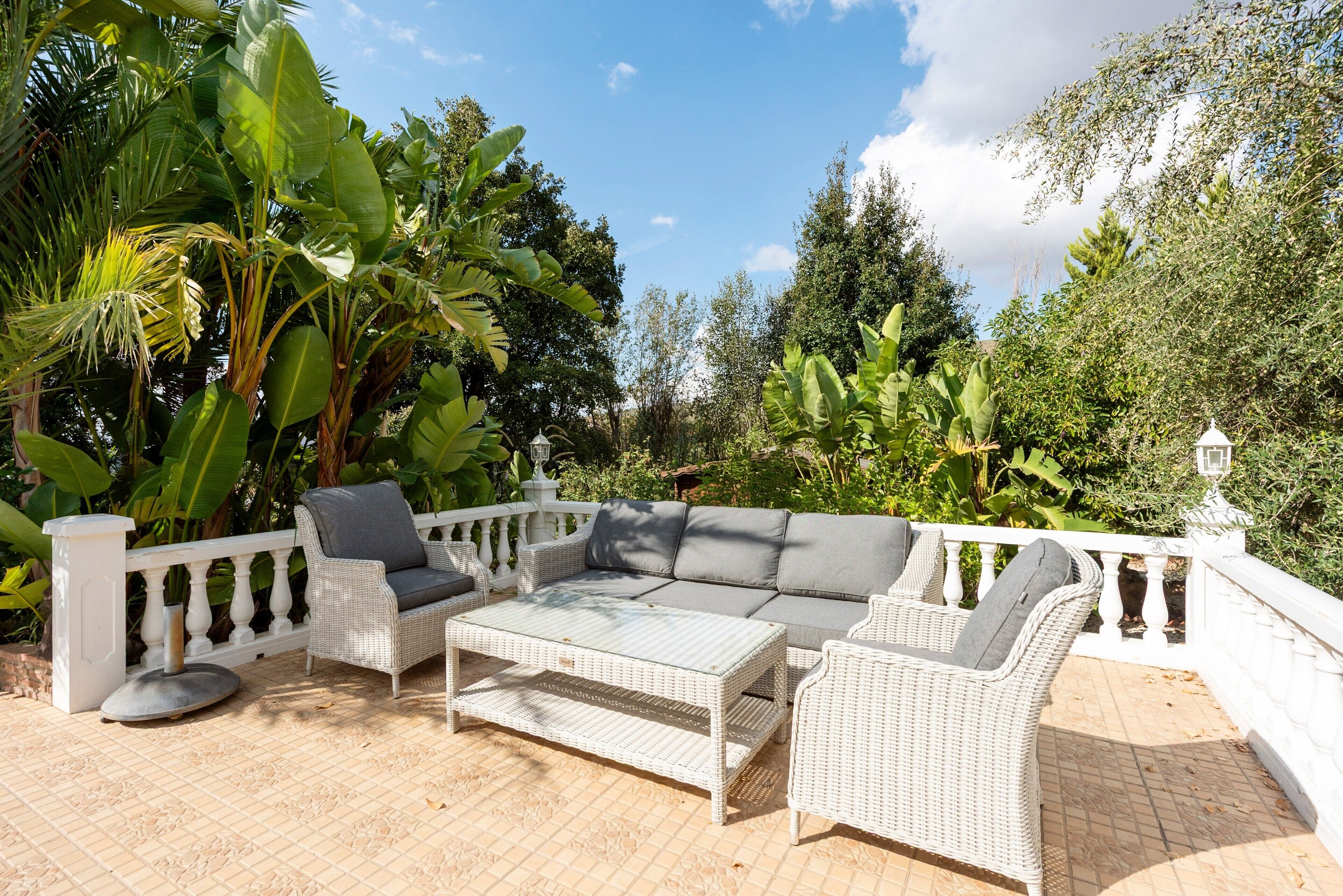 An outdoor patio with wicker furniture, a white railing, and lush greenery in the background.