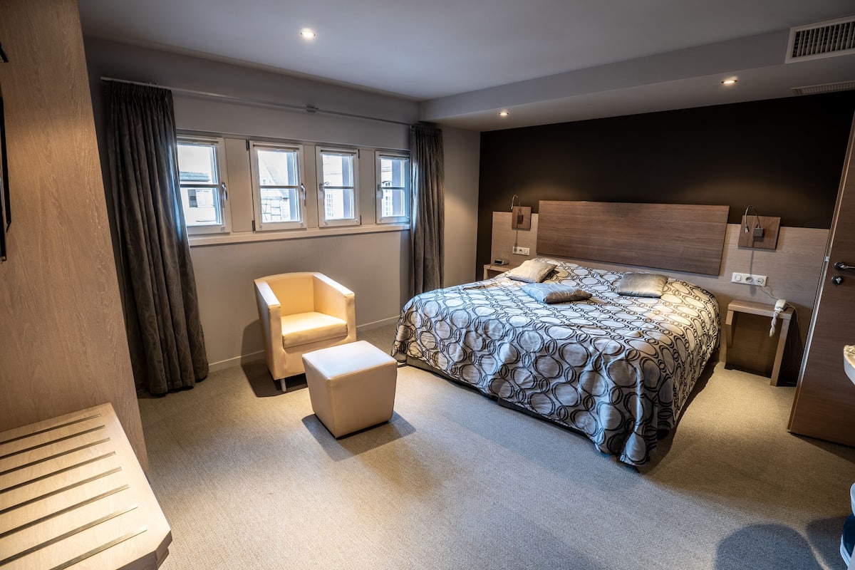 A modern hotel room with a large bed, a chair, a small table, and a window with curtains.
