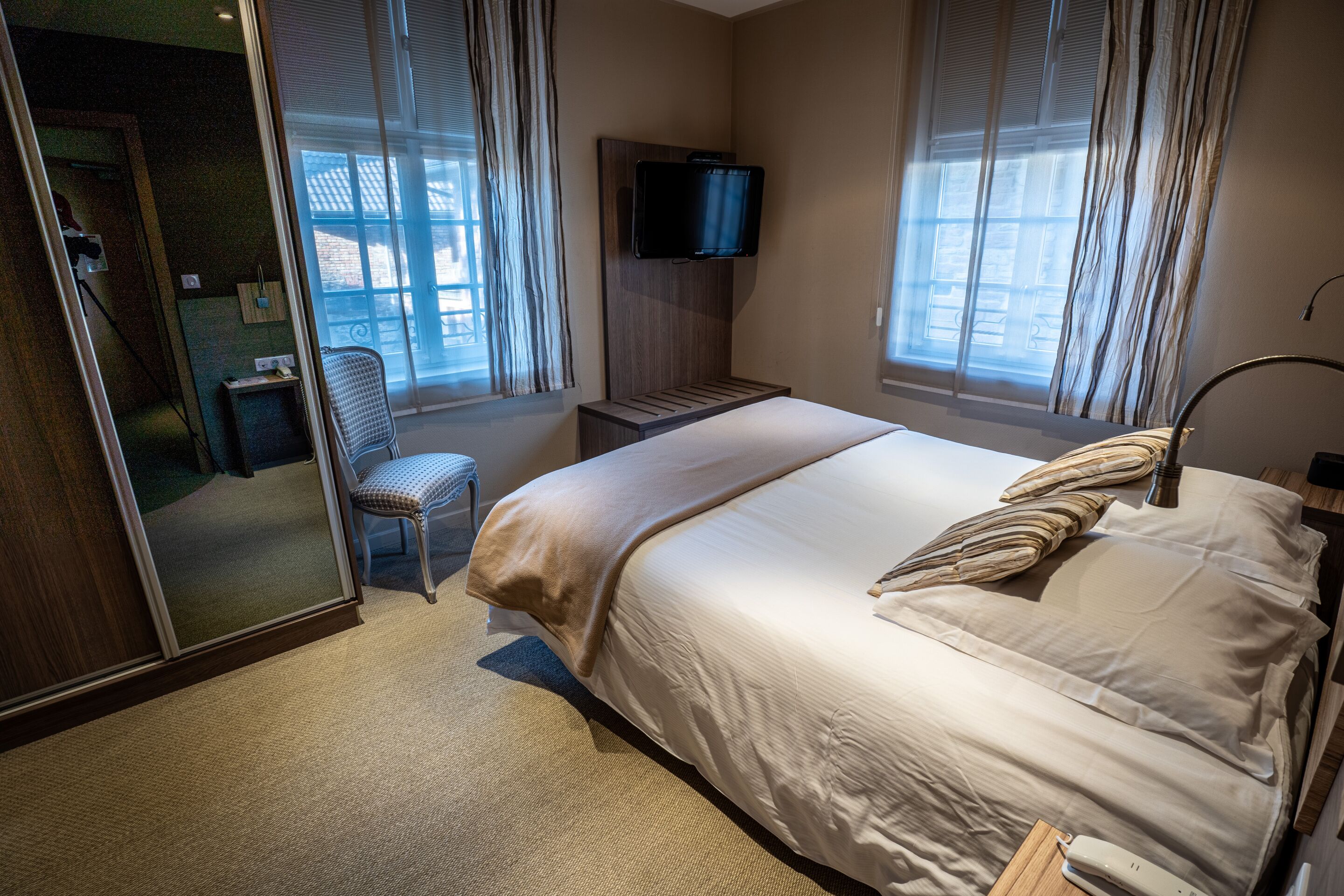 A hotel room with a bed, a TV mounted on the wall, a chair, and a window with curtains.