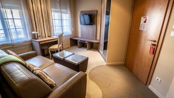 A hotel room with a leather sofa, a small desk with a chair, a television mounted on a wooden panel, and a door with a fire alarm.