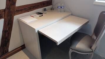 A small, white desk with a chair, a remote control, two water bottles, and a power outlet on the wall.