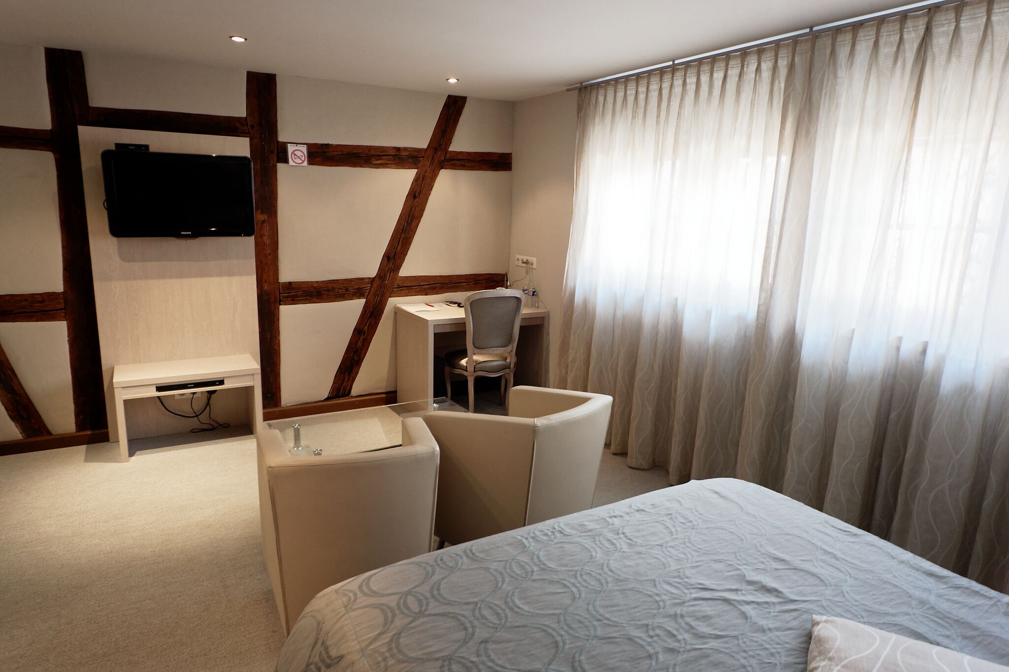 Una habitación de hotel con una cama, un escritorio con silla, un televisor montado en la pared y una ventana con cortinas.