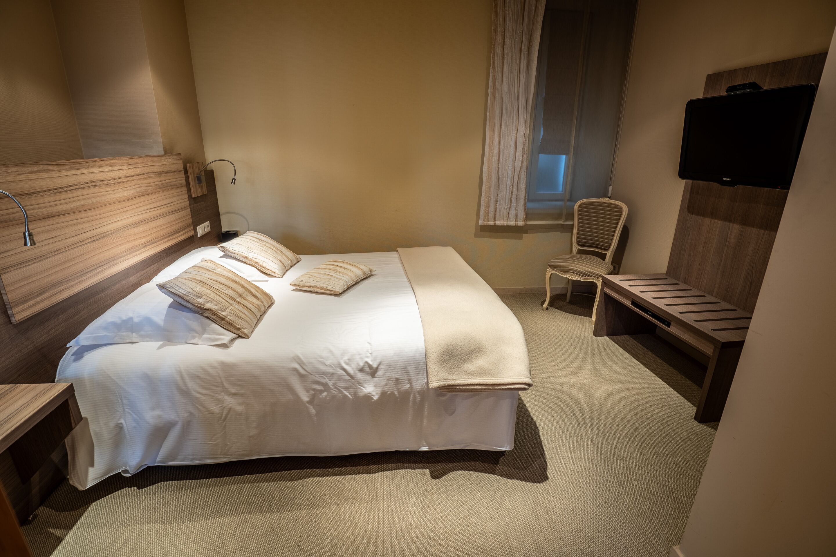 Ein Hotelzimmer mit einem großen Bett, einem Nachttisch, einem Sessel, einem Fernseher und einem Fenster mit Vorhängen.