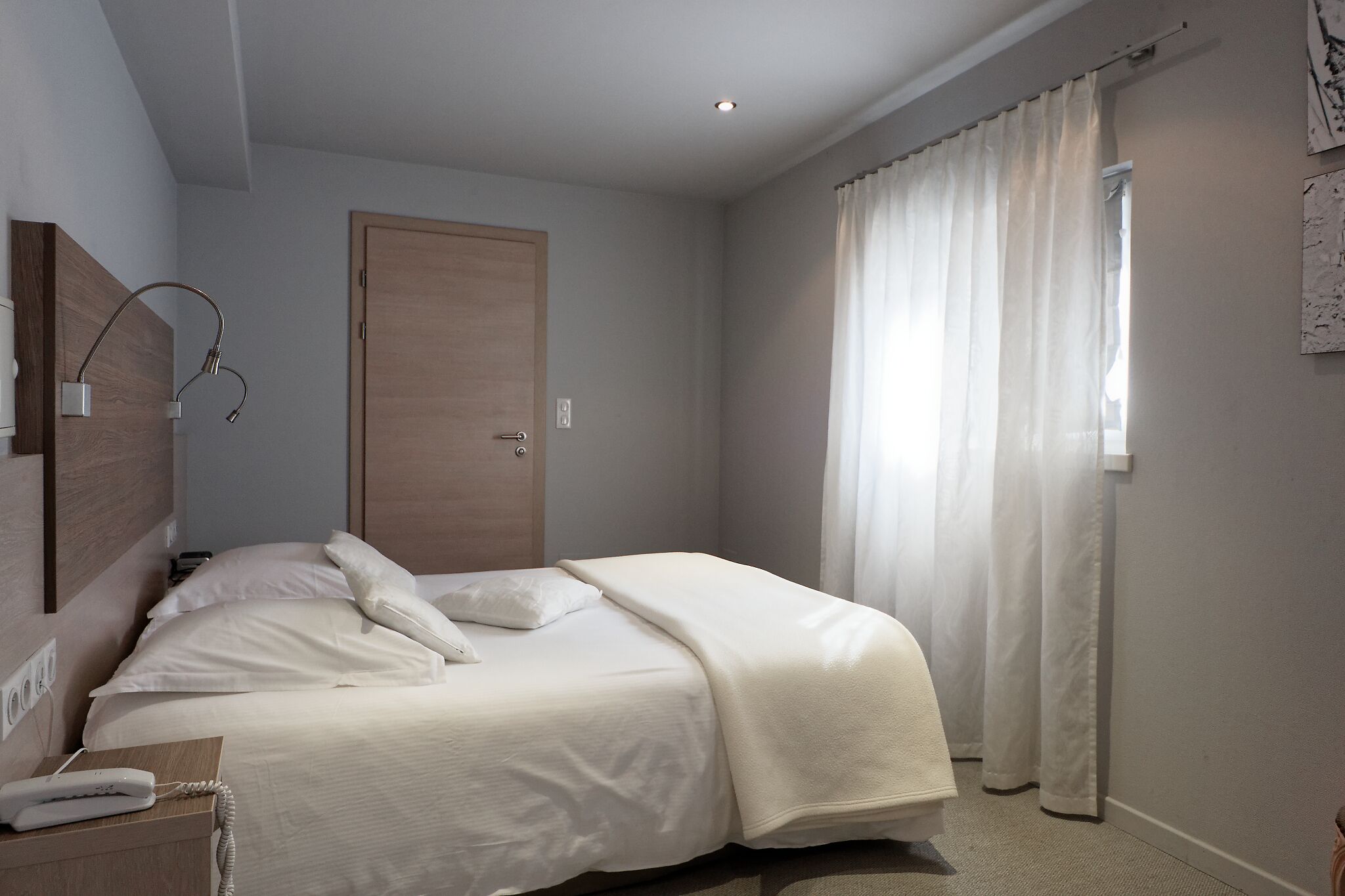 A neatly arranged bedroom with a bed, a bedside table with a phone, a wooden door, and a window with curtains.