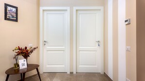 Un pasillo con dos puertas blancas, una pequeña mesa con un jarrón de flores secas y un cuadro en la pared.