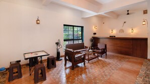 A spacious living area with a wooden sofa, a small table, and a fan. There is a window with a view of greenery outside. - Vaarsa By Bay Hotels (Nagaon)